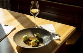 Teller mit Fleisch, Knödeln und Soße, Weinglas auf Holztisch im Sonnenlicht.