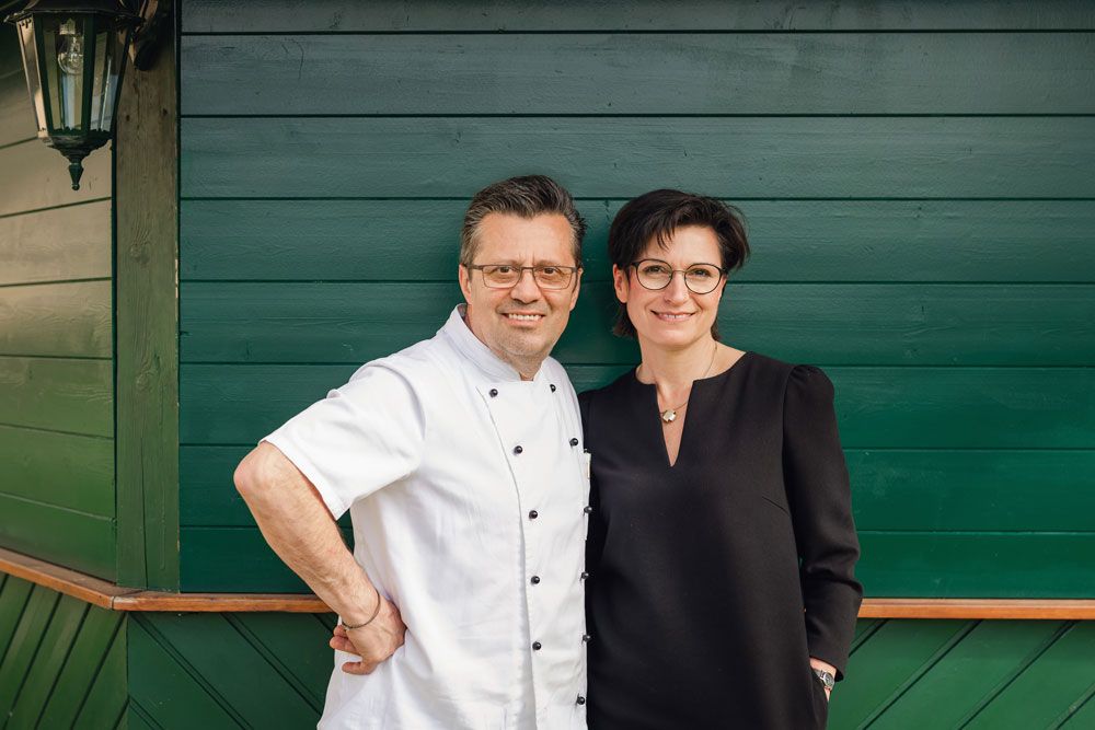 Ein Mann in Kochjacke und eine Frau in schwarzem Kleid stehen vor einer grünen Holzwand.